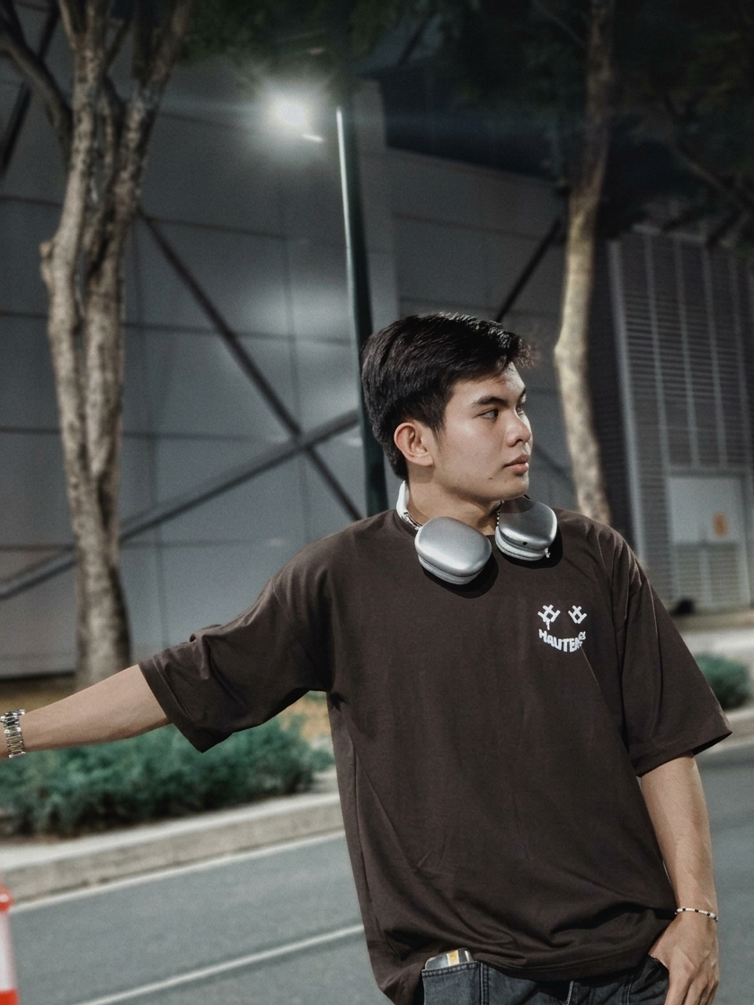 Man wearing a brown t-shirt with a logo, standing outdoors near trees and a building.